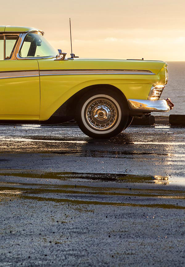 A yellow Ford Fairlane parked on a wet pavement with a sunset in the background.