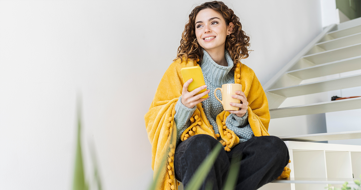 Women sitting on staircase in a yellow blanket, holding phone and a cup of tea