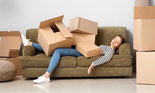 A woman, tired from moving, laying on the couch with her eyes closed covered in boxes