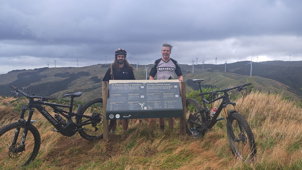 Two mountain bikers with their bikes. Wind-farm in the background