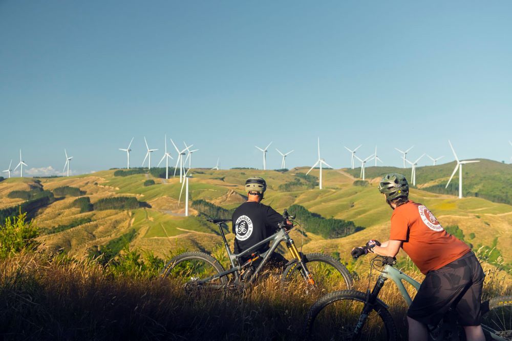 Two mountain-bikers with backs to camera looking at view of Turitea wind farm